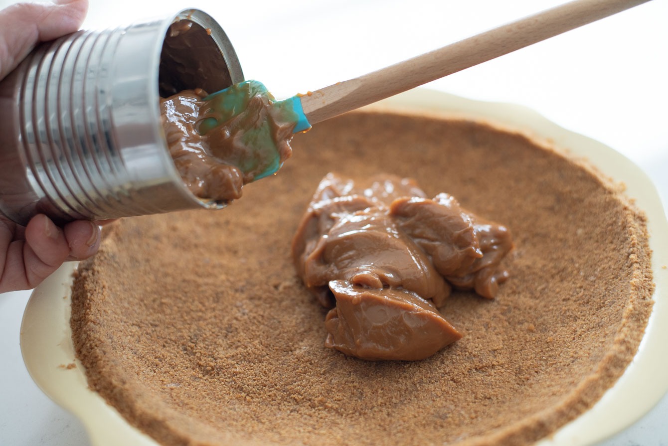 A spatula is taking dulce de leche from the can and putting on to a graham cracker pie crust.
