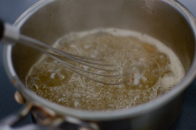 A whisk is stirring the vigorously boiling honey syrup in a pan.