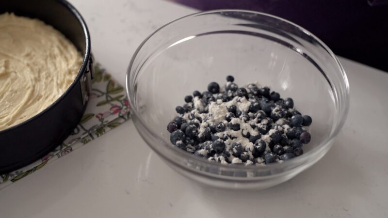 A spoonful of flour is added to the fresh blueberries in a bowl.