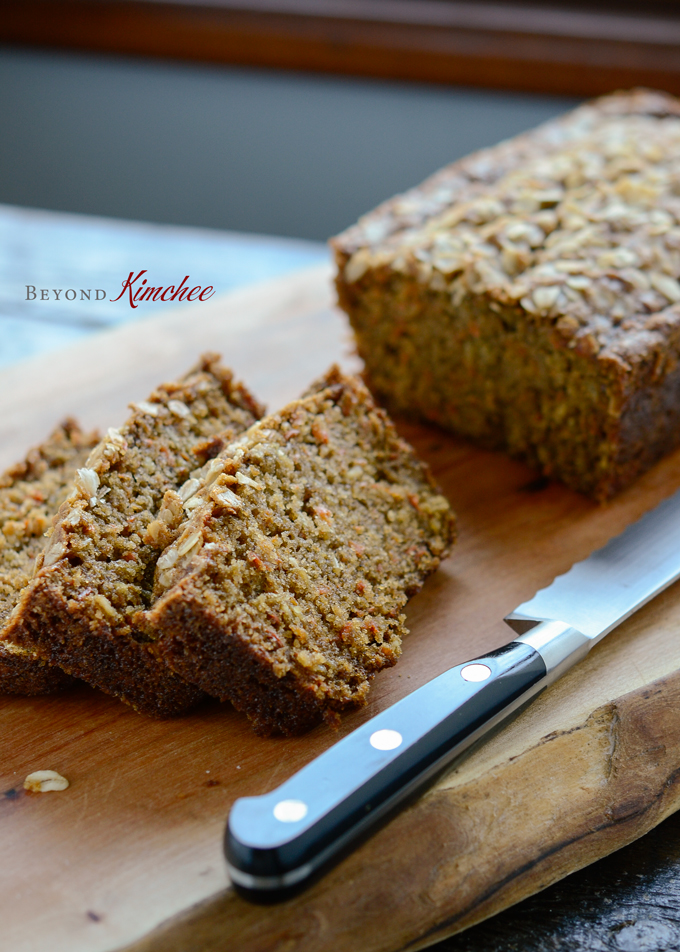 A loaf of carrot oat bread bread is sliced with a knife on a cutting board.