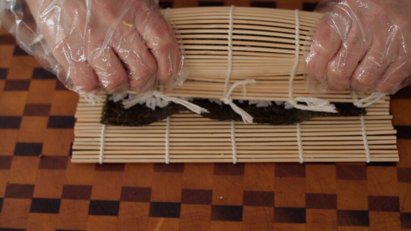 Hands are squeezing the bamboo rolling mat lightly after it is rolled to secure gimbap inside.