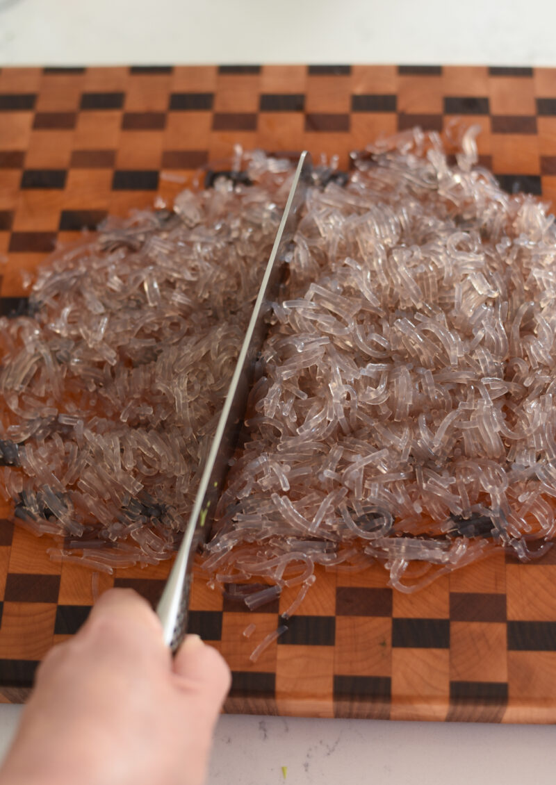 Boiled Korean glass noodles are chopped to small on a cutting board with a knife.