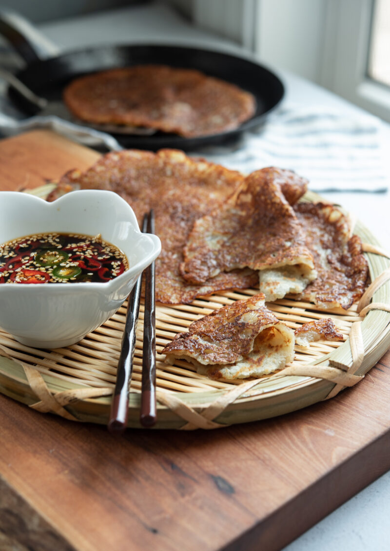Thin potato pancakes are served on a woven wood plate with a bowl of soy chili dipping sauce.