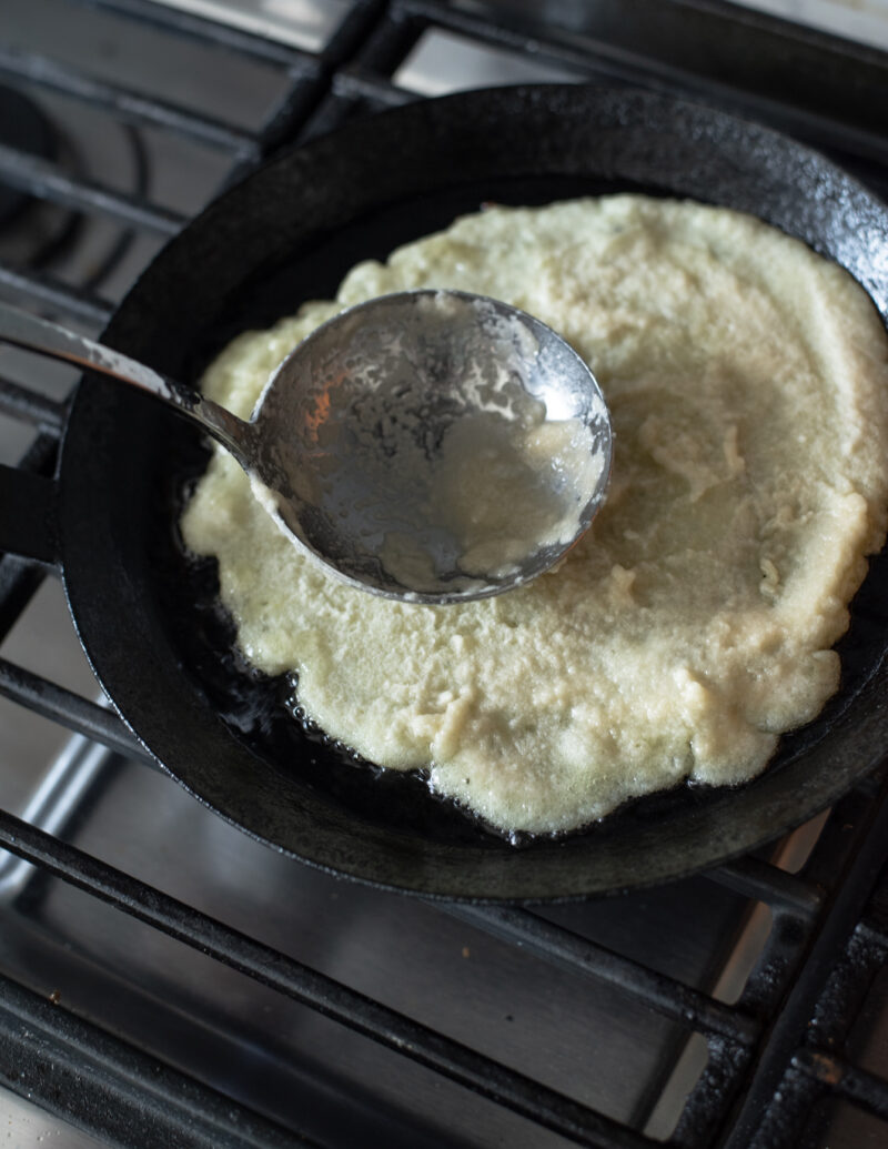 A ladle is spreading the potato pancake batter in a skillet to smooth out the top.