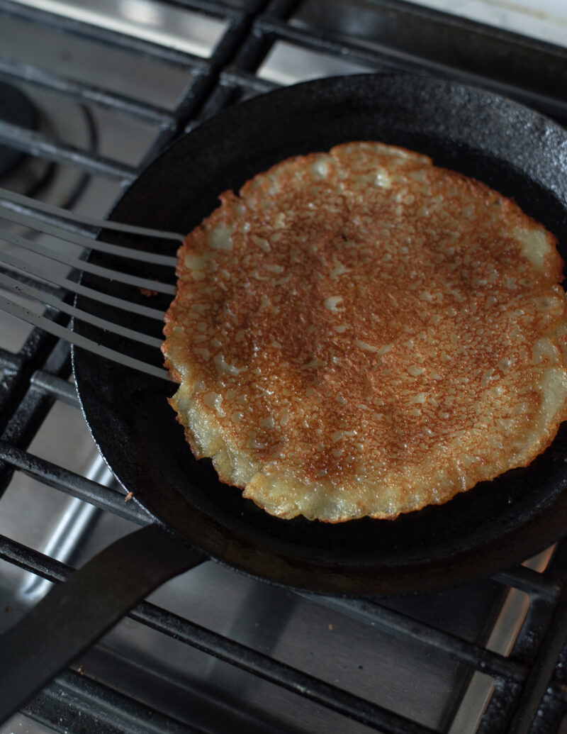 A spatula is turning the potato pancake to the other side revealing the golden brown color of pancake.