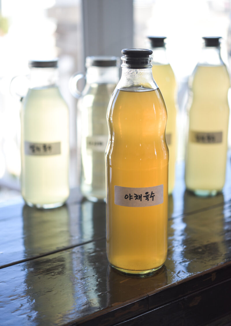 Vegetable stock is showing its golden hue through a glass bottle with a Korean label attached.