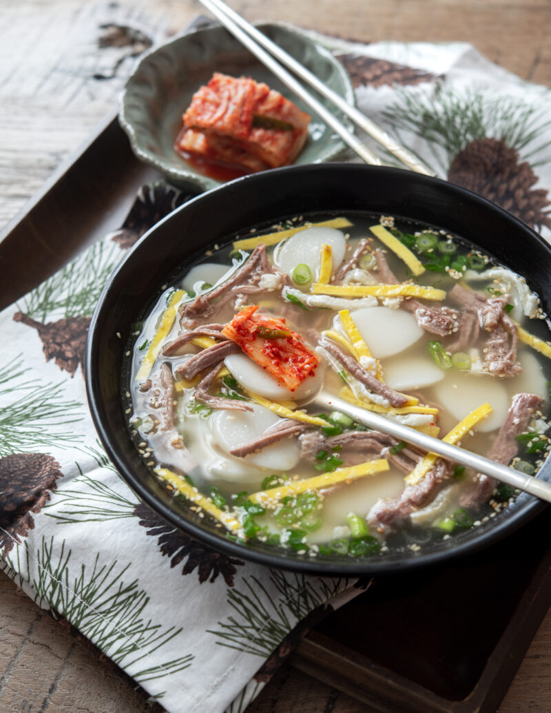 Korean rice cake soup in a clear broth with beef and egg is served with kimchi.