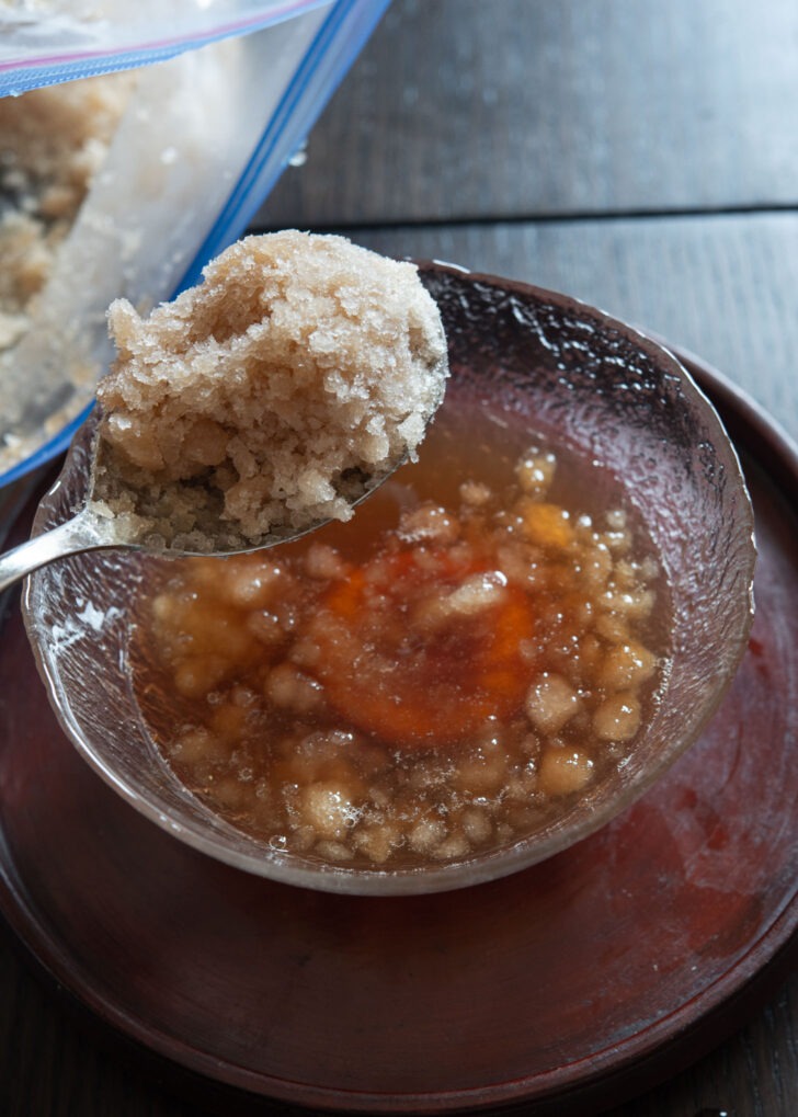 A spoonful of crushed cinnamon ice is added to sujeonggwa in a bowl.